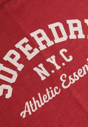 Rode katoenen t-shirt met witte grafische tekst "SUPERDRY NYC Athletic Essentials" in vetgedrukte en cursieve lettertypen. Zachte textuur, casual ontwerp.