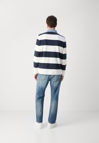 Tamsiai mėlynas ir baltas dryžuotas polo marškinėlis ilgomis rankovėmis, derinamas su šviesiai mėlynais džinsais ir baltais sportbačiais, matomas iš nugaros, demonstruojantis kasdienį aprangos stilių.