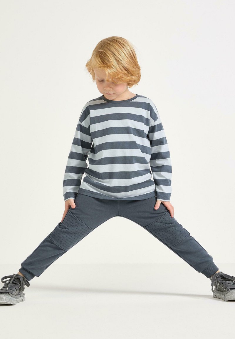 Bambino con capelli biondi che indossa una maglietta a maniche lunghe grigia a righe e pantaloni scuri, in piedi con le gambe larghe su uno sfondo bianco uniforme.
