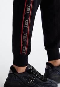 Schwarze Jogginghose mit einem roten seitlichen Streifen und einem dekorativen Muster. Das Modell trägt schwarze Sneaker, die mit Strasssteinen verziert sind.