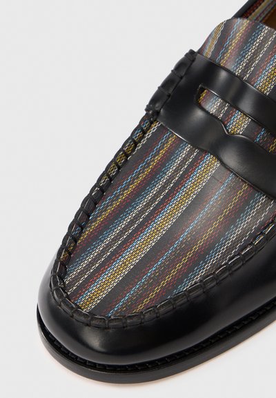 Fekete bőr mokaszin színes csíkokkal díszített textil felsőrésszel, piros, kék és sárga árnyalatokkal sötét háttér előtt.