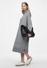 Γκρι πλεχτό φόρεμα με ψηλό λαιμό και ριμπέ επένδυση, πλαϊνές σχισμές, συνδυασμένο με μία μεγάλη μαύρη τσάντα ώμου με υφή και λευκά αθλητικά παπούτσια.