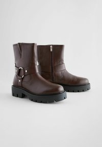 Botas de tobillo de cuero marrón con un acabado texturizado, cierre lateral con cremallera, tira decorativa con hebilla y suelas de goma negras gruesas.