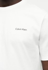 Λευκό πουκάμισο με κοντά μανίκια και μικρό μαύρο λογότυπο "Calvin Klein" στο αριστερό στήθος που φοριέται από άτομο με σκουρόχρωμο δέρμα.