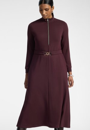 Femme portant une robe longue à manches longues, col montant, de couleur violet foncé avec une fermeture éclair à l'avant et une ceinture à boucle dorée à la taille.