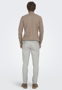 Suéter de punto marrón claro con cuello alto, combinado con pantalones gris claro con bolsillos traseros y zapatos en tonos neutros.