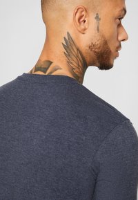 Marineblaues, tailliertes T-Shirt mit rundem Halsausschnitt, strukturiertem Stoff und sichtbaren Schulternähten; Tätowierungen am Hals umfassen Flügel- und Dolchdesigns.