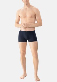 Barberløsf mand står barfodet iført marineblå tætsiddende boxershorts, hænderne løst foldet foran sig, på en lys ensfarvet baggrund.
