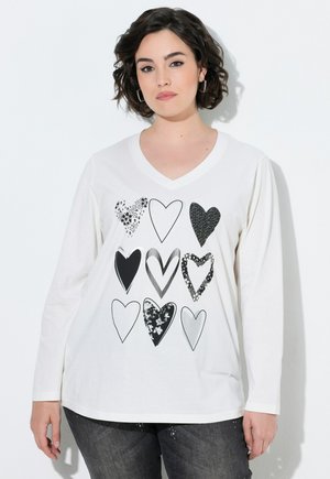 Femme aux cheveux courts et foncés portant une chemise blanche à manches longues avec neuf motifs de cœurs en noir et blanc, debout sur un fond blanc.