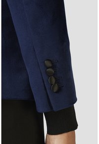 Le maniche della giacca blu navy presentano un tessuto liscio con quattro bottoni neri e una texture a coste nera al polsino. Design pulito con vestibilità sartoriale.