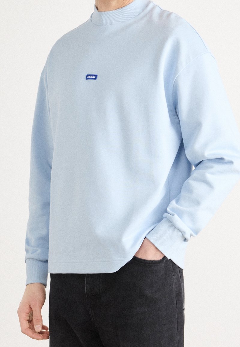 Homme portant un sweat-shirt bleu clair à manches longues avec le logo "HUGO" sur la poitrine et un pantalon noir, main partiellement dans la poche.