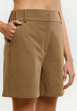 Beige op maat gemaakte korte broek met ceintuurlussen en zijzakken, gedragen door een persoon met één hand in de zak en de andere ontspannen langs het lichaam.