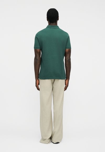 Homme debout, de dos, portant un polo à manches courtes vert foncé et un pantalon large beige, sur fond blanc uni.