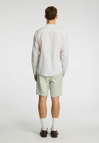 Camisa blanca de manga larga, pantalones cortos verde claro, zapatos marrones, calcetines blancos. Textura sutil en la camisa, cuello y diseño de corte estándar.
