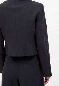 Czarna żakiet skrojony na miarę o skróconym fasonie, gładkiej fakturze, usztywnionych ramionach i skośnych kieszeniach z tyłu. Widoczne przeszycia.