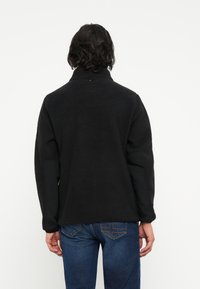 Svart fleecejakke med høy krage, teksturert stoff og kontrasterende svarte ermer. Brukt med blå jeans med dekorativ søm.
