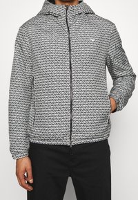 Svart och vit mönstrad jacka med huva, med en framdragkedja och sidofickor. Slät textur och modern geometrisk design.