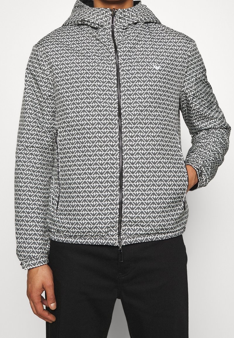 Svart och vit mönstrad jacka med huva, med en framdragkedja och sidofickor. Slät textur och modern geometrisk design.