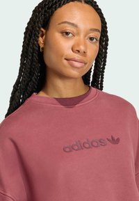 Felpa bordeaux con scollo a girocollo a coste e logo "adidas" in rilievo sul davanti. Texture liscia, vestibilità rilassata e design minimale.