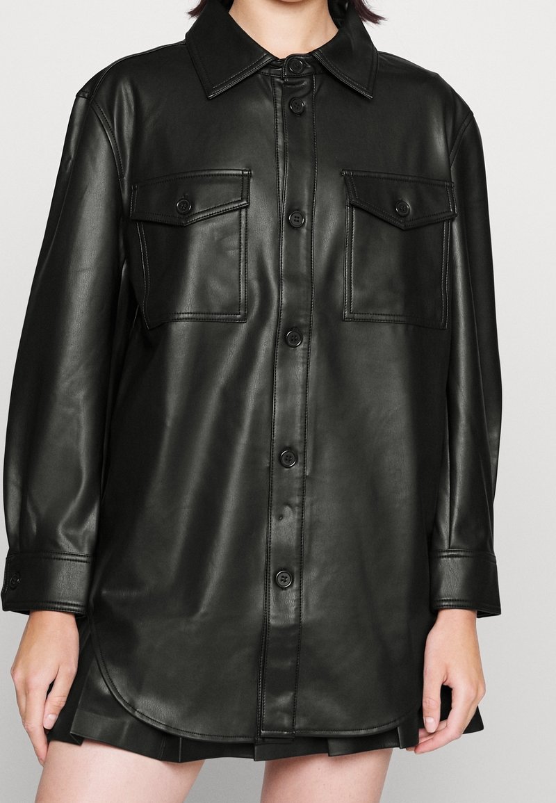 Černá koženka vycházející z košile s límcem, dvěma náprsními kapsami a zapínáním na knoflíky, s zakřiveným lemem a dlouhými rukávy.