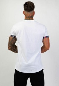 Vit bomullströja med korta ärmar som har ett blommönster vid ärmsluten. På baksidan finns en tryckt etikett som står "LIMITÉ" nära fållen.