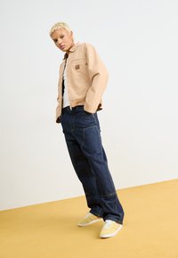 Beige zip-up jacket with chest pocket over a black top, paired with loose blue jeans and yellow sneakers on a yellow floor.