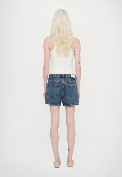 Shorts en denim avec un ourlet effiloché, délavage bleu, deux poches arrière et une étiquette de taille blanche. Associés à un débardeur blanc ajusté.