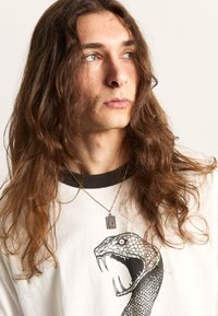 Camisa blanca de manga larga con un gráfico de serpiente negra, con un cuello redondo negro y un collar plateado en capas. Tela suave, corte holgado.