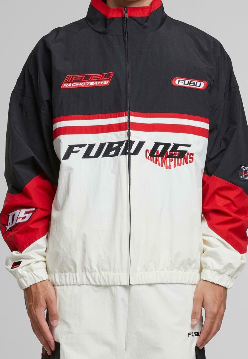 Homme portant une veste de course FUBU noire, rouge et blanche avec logos et texte, fermée devant, les mains le long du corps.