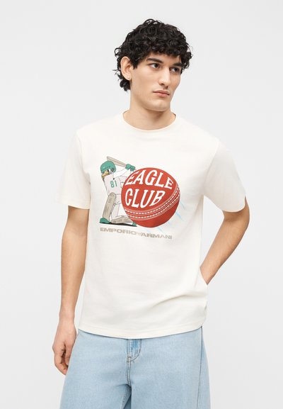 Νεαρός άνδρας με ανοιχτόχρωμο τζιν, φοράει λευκό μπλουζάκι με μια εικονογράφηση παίκτη κρίκετ και το κείμενο "Eagle Club" στο μπροστινό μέρος.