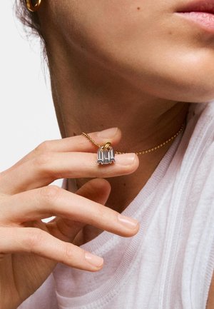 Persona que lleva un collar de cadena dorada con un colgante plateado con la palabra "MOM", sosteniendo el colgante entre el pulgar y el índice, vistiendo camisa blanca y pendiente de aro dorado.