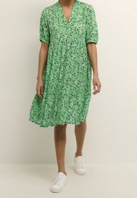 Robe florale verte avec un col en V, des manches courtes bouffantes et une jupe à étages. Portée avec des baskets blanches. Caractérisée par un tissu léger et texturé.
