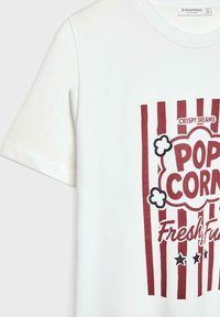 T-shirt en coton blanc avec un graphique de popcorn rouge et des rayures, texte "Crispy Dreams" et étoiles, à manches courtes, coupe décontractée.