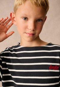 Fekete-fehér csíkos póló kerek nyakkivágással és rövid ujjakkal, amelyen piros "Superdry" logó található a mellrészen.