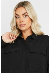 Zwarte button-up shirt met een gestructureerde stof, twee voorzakken en een kraag, gedragen door een persoon met lang, golvend haar.