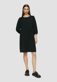 Schwarzes Kleid mit entspannter Silhouette, rundem Ausschnitt und Puffärmeln. Getragen mit schwarzen Slippers, die eine glatte Textur zeigen.