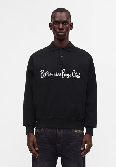 Мъж, облечен в черна поло риза с дълъг ръкав с бял текст "Billionaire Boys Club", комбинирана с тъмносини дънки, стои пред едноцветен фон.