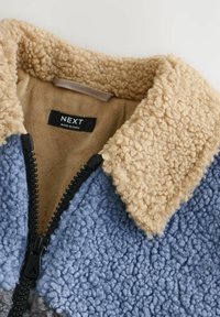 Veste en polaire bleu clair avec un col texturé crème. Elle présente une fermeture éclair noire et une étiquette intérieure avec le nom de marque "NEXT."