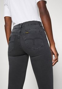 Dark grey high-waisted jeans with a fitted shape, featuring two back pockets, yellow logo accent, and contrast stitching along the seams.