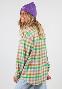 Chemise en flanelle à motif carreaux verts, violets, marron et crème ; manches longues ; ourlet arrondi ; présente un petit logo vert sur l'épaule.