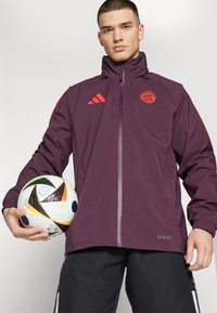 Giacca leggera impermeabile color marsala con zip frontale e cappuccio; presenta dettagli rossi e un logo; abbinata a pantaloni neri e a un pallone da calcio multicolore.
