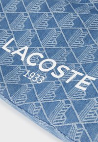 Džinsinis maišelis su šviesiai mėlynu fonu, išdėstytas pakartotiniu raštu ir ryškiu baltu tekstu "LACOSTE 1933" apačioje.