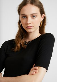Schwarzes, eng anliegendes Oberteil mit kurzen Ärmeln. Das Modell trägt goldene Creolen und geschichtete Halsketten mit rechteckigen Anhängern. Schlichtes Design und Textur.