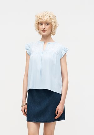 Szőke nő göndör hajjal, világoskék ujjatlan fodros vállú blúzzal és sötétkék farmer szoknyával, fehér háttér előtt áll.