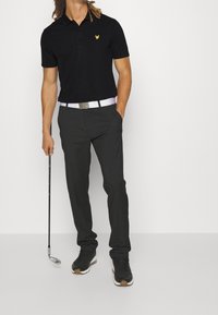 Mężczyzna w czarnej koszulce polo i ciemnych spodniach trzymający kij golfowy, stojący na jednolitym białym tle.