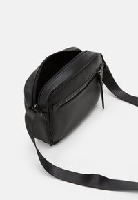 Borsa a tracolla nera in pelle con finitura testurizzata, scomparto principale con zip e tasca frontale piatta con accentuazione del logo. Cinghia regolabile.