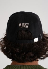 Svart baseballcaps med brodert "MODERN YUPPIES" i beige bak, med en metallrem for justering og en strukturert design.