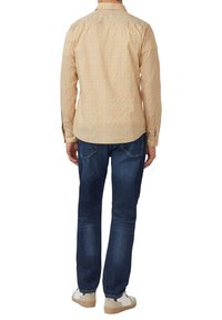 Heren overhemd met lange mouwen in mosterdkleur met een bloemenpatroon. Gecombineerd met donkerblauwe spijkerbroek en witte sneakers. Achteraanzicht toont de pasvorm van het overhemd.