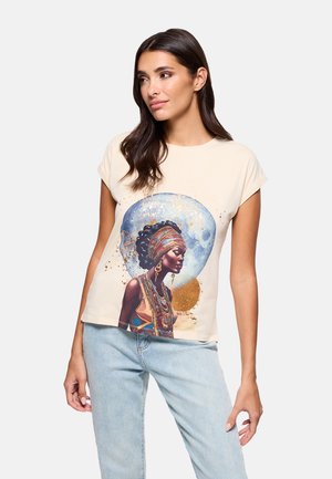 Mujer que lleva una camiseta beige con una impresión artística colorida de una mujer con vestimenta y joyas tradicionales africanas, combinada con vaqueros azul claro.