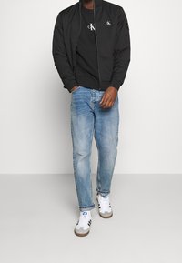 Giacca bomber nera sopra una maglietta nera con logo, abbinata a jeans azzurri chiari e sneakers grigie con strisce nere e dettagli bianchi.